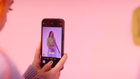 Studio-Shot-Of-Woman-Taking-Photo-Of-Friend-Dancing-On-Mobile-Phone-Against-Pink-Background